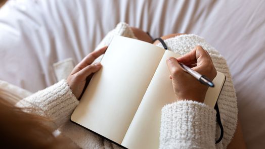 Một người phụ nữ đang cầm một cuốn sổ tay v&agrave; một c&acirc;y b&uacute;t, chuẩn bị bắt đầu viết nhật k&yacute; tr&ecirc;n giường